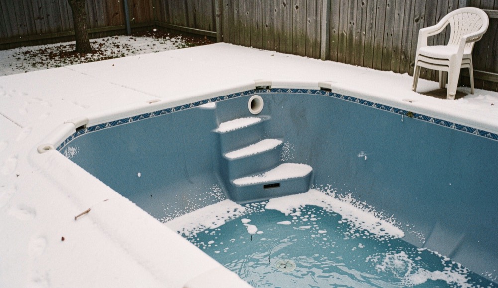 Corner of a blue vinyl-lined pool with steps and a skimmer opening under a light dusting of first snow, stacked garden chairs nearby